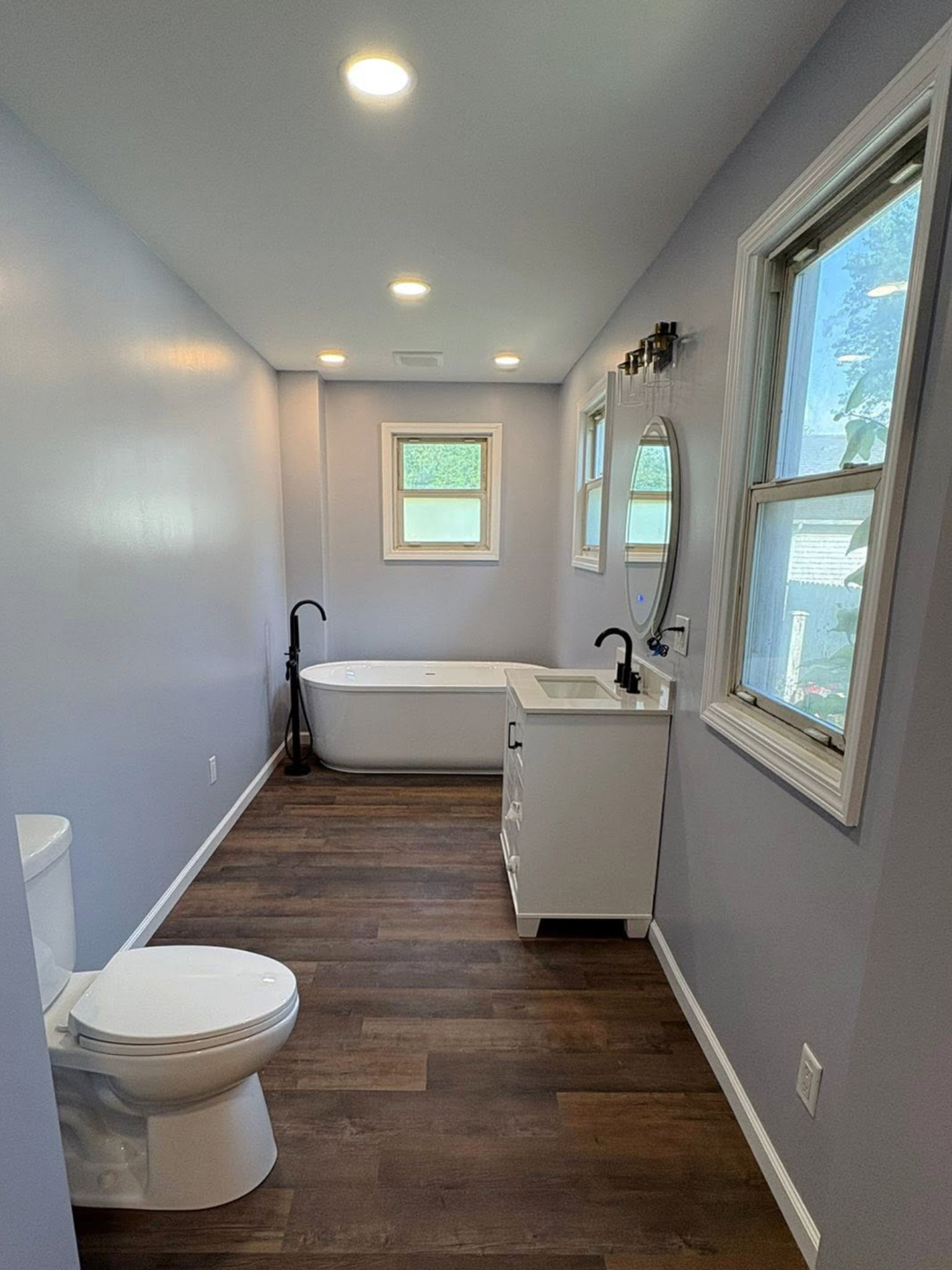 A narrow bathroom with a freestanding tub, new vanity and dark plank flooring.